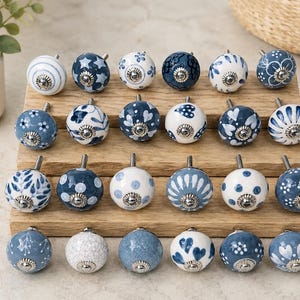 Pu&ograve; includere: Una collezione di ventiquattro pomelli rotondi in ceramica per mobili con vari motivi blu e bianchi, tra cui strisce, stelle, pois, motivi floreali e cuori. Ogni pomello ha una base in metallo color argento e una vite centrale.