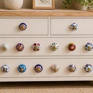 May include: A cream-colored wooden dresser with a light wood top, featuring four small upper drawers and two large lower drawers. The drawers are fitted with fourteen round, multi-colored ceramic knobs, each displaying unique floral patterns.