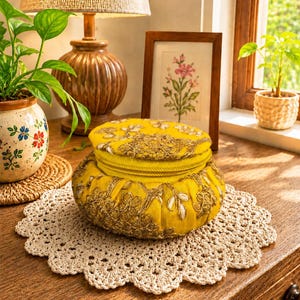 May include: A round, yellow fabric storage container featuring intricate gold-colored embroidery and a zippered lid. The container has a pleated side design and sits on a circular, cream-colored crocheted doily with a scalloped edge.