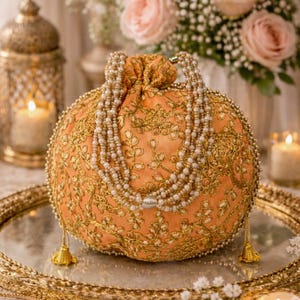 Puede incluir: Un bolso de mano bordado en color naranja con detalles dorados y una correa de cuentas. El bolso redondo se exhibe sobre un espejo con borde dorado, con flores y velas en el fondo, creando una estética elegante.