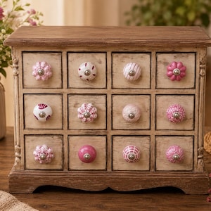 Pode incluir: Um baú de armazenamento de madeira com doze gavetas quadradas, cada uma com um puxador de cerâmica decorativo exclusivo em vários tons de rosa e branco. O armário tem um acabamento castanho claro desgastado com topo e base mais escuros.
