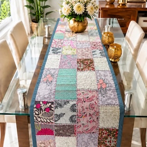 Könnte beinhalten: Ein rechteckiger Tischläufer mit einem Patchwork-Design aus verschiedenen Stoffquadraten in Rosa-, Türkis-, Grau- und Weißtönen. Der Läufer hat einen einfarbigen blauen Rand und durchgehend sichtbare Steppnähte, flach auf einem Glastisch.