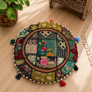Puede incluir: Un cojín de suelo redondo con un diseño de patchwork de varias telas en tonos verde, rojo, dorado y negro. La superficie está decorada con intrincados bordados florales, detalles de hilo metálico y pequeños pompones blancos en el borde.