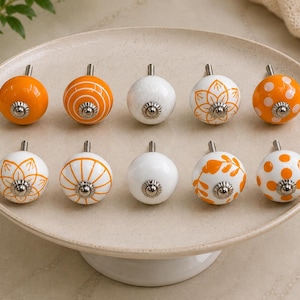 May include: A collection of ten decorative ceramic cabinet knobs. The knobs are round with a silver metal stem. Some are white with orange floral designs, others are solid orange or white with orange polka dots. They are displayed on a white oval platter.