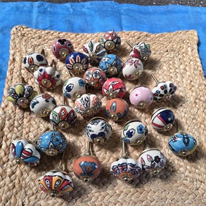 May include: A collection of round ceramic cabinet knobs featuring various hand-painted floral and geometric patterns in colours including blue, red, pink, orange, yellow, and green. Each knob has a metallic base and a central screw attachment.