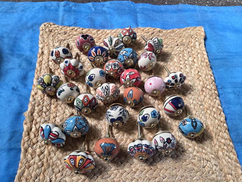 May include: A collection of round ceramic cabinet knobs featuring various hand-painted floral and geometric patterns in colours including blue, red, pink, orange, yellow, and green. Each knob has a metallic base and a central screw attachment.
