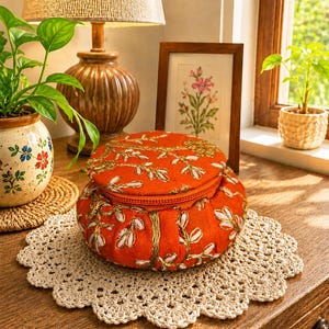May include: A round, orange fabric storage box with a zippered lid, featuring intricate gold and white floral embroidery. The container has a soft, padded structure and sits on a circular, cream-colored crocheted lace doily.