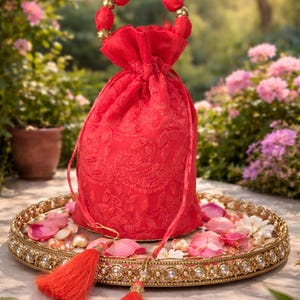 Peut inclure: Une pochette à cordon rouge vif ornée de broderies ton sur ton complexes et d'une poignée circulaire agrémentée de sphères en tissu rouge et de perles dorées. Le sac comporte deux longs pompons rouges suspendus au cordon de serrage.