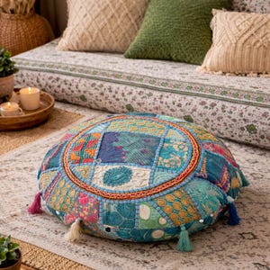 May include: A round floor cushion featuring a vibrant patchwork design with various shades of blue, green, and pink fabric panels. The cushion is decorated with intricate embroidery, gold-toned patterns, and small tassels along the outer edge.