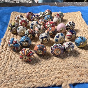 May include: A collection of round ceramic cabinet knobs featuring various colourful hand-painted floral, geometric, and striped patterns. Each knob has a central gold-toned metal hardware piece and a smooth, glossy finish with diverse designs.