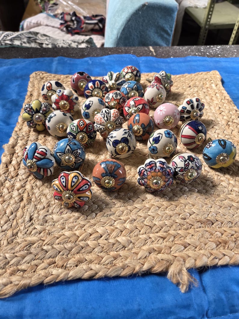 May include: A collection of round ceramic cabinet knobs featuring various colourful hand-painted floral, geometric, and striped patterns. Each knob has a central gold-toned metal hardware piece and a smooth, glossy finish with diverse designs.