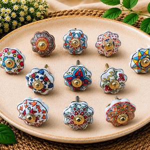 Op de afbeelding: Een verzameling van tien ronde keramische ladeknoppen met geschulpte randen, elk met een uniek veelkleurig bloemen- of geometrisch patroon en een centrale goudkleurige metalen schroefkop, gerangschikt op een lichtbeige ronde schaal.