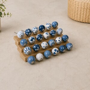 Pu&ograve; includere: Una collezione di ventiquattro pomelli rotondi in ceramica per mobili disposti su un espositore in legno a gradini. Ogni pomello presenta un motivo unico in blu e bianco, inclusi disegni floreali, a pois, a cuore e a righe con finiture.