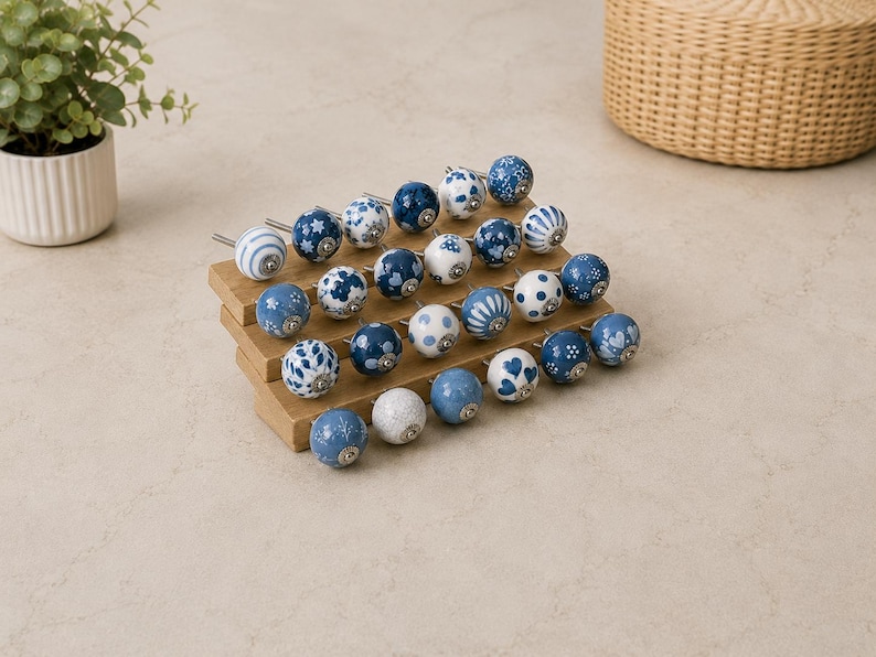 Pu&ograve; includere: Una collezione di ventiquattro pomelli rotondi in ceramica per mobili disposti su un espositore in legno a gradini. Ogni pomello presenta un motivo unico in blu e bianco, inclusi disegni floreali, a pois, a cuore e a righe con finiture.
