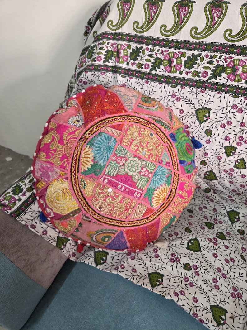 May include: A round, multi-colored patchwork floor cushion featuring intricate embroidery, small circular mirrors, and vibrant fabric panels in shades of pink, orange, yellow, and blue, finished with small decorative tassels along the outer edge.