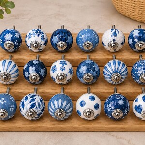 Pu&ograve; includere: Diciotto pomelli rotondi in ceramica per mobili disposti su tre file su un espositore in legno. Ogni pomello presenta un motivo unico in blu e bianco, inclusi disegni floreali, a righe, a pois, a stelle e a cuori con finiture argentate.
