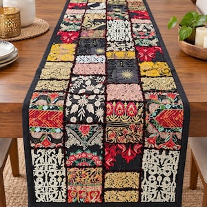 Puede incluir: Un camino de mesa rectangular con un diseño de retazos de varios cuadrados de tela. El camino muestra una mezcla de textiles negros, rojos, amarillos y blancos con bordados intrincados, encaje y patrones florales, acabado con un borde negro.