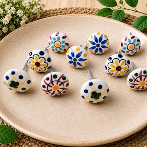 May include: A collection of nine round ceramic knobs featuring various hand-painted floral and geometric patterns in blue, yellow, green, and red colours. Each knob has a silver-toned metal screw extending from the back for mounting purposes.
