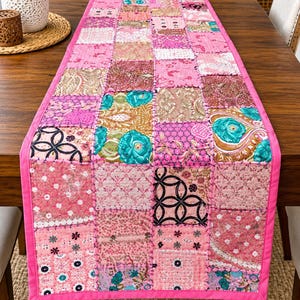 Puede incluir: Un camino de mesa rectangular con un diseño de patchwork de varios cuadrados de tela en tonos rosa, turquesa y marrón. El camino está acolchado con costuras visibles y acabado con un borde de tela rosa brillante sólido en los extremos.