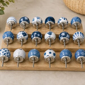 Pu&ograve; includere: Una collezione di diciotto pomelli rotondi in ceramica per mobili disposti su tre file su un espositore in legno. Ogni pomello presenta un motivo unico in blu e bianco, inclusi disegni floreali, a pois, a righe e geometrici.