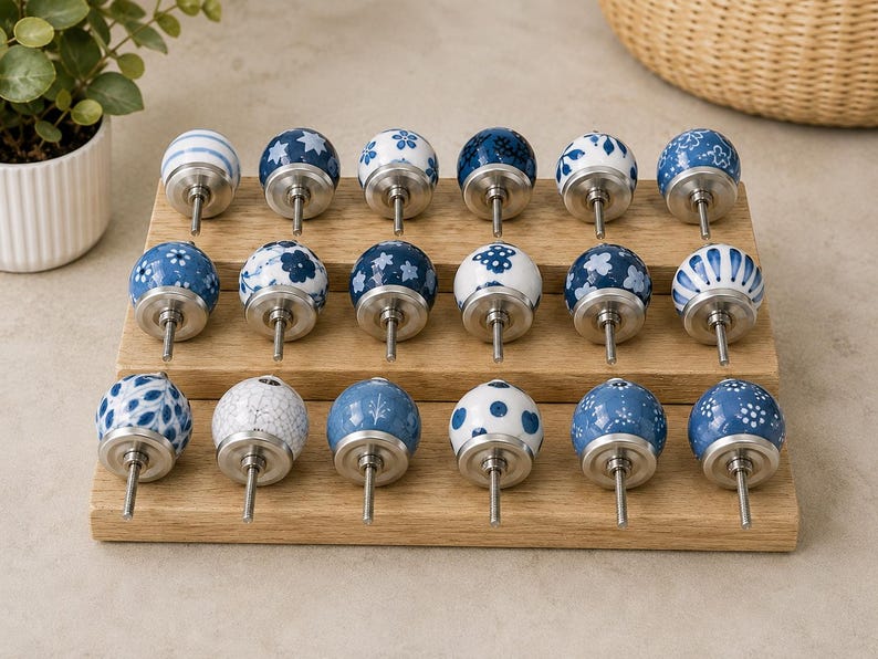 Pu&ograve; includere: Una collezione di diciotto pomelli rotondi in ceramica per mobili disposti su tre file su un espositore in legno. Ogni pomello presenta un motivo unico in blu e bianco, inclusi disegni floreali, a pois, a righe e geometrici.
