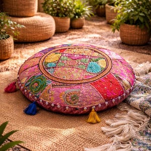 Könnte beinhalten: Ein runder, pinkfarbener Bodenkissen mit farbenfrohen Patchwork- und Stickdetails. Das Kissen hat einen goldenen Rand und ist mit Quasten in verschiedenen Farben verziert. Es liegt auf einem gewebten Teppich, was auf einen Boho- oder globalen Einrichtungsstil hindeutet.