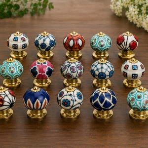 May include: A collection of fifteen decorative ceramic cabinet knobs. Each knob features a unique hand-painted design in various colors, including blue, red, and white, with a gold-toned base. The knobs are arranged on a wooden surface.