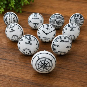 May include: A collection of ten round white ceramic knobs featuring various black printed designs, including clock faces, compasses, and vintage-style typography such as Paris Clocks, London Clocks, Suave Absinthe, and Olive Oil Soft Soap.