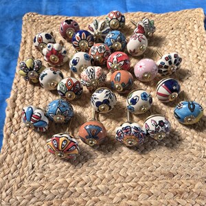 May include: A collection of twenty-four round ceramic cabinet knobs featuring various hand-painted floral and geometric patterns in colours including blue, red, orange, pink, and white, each finished with a central gold-toned metal hardware piece.