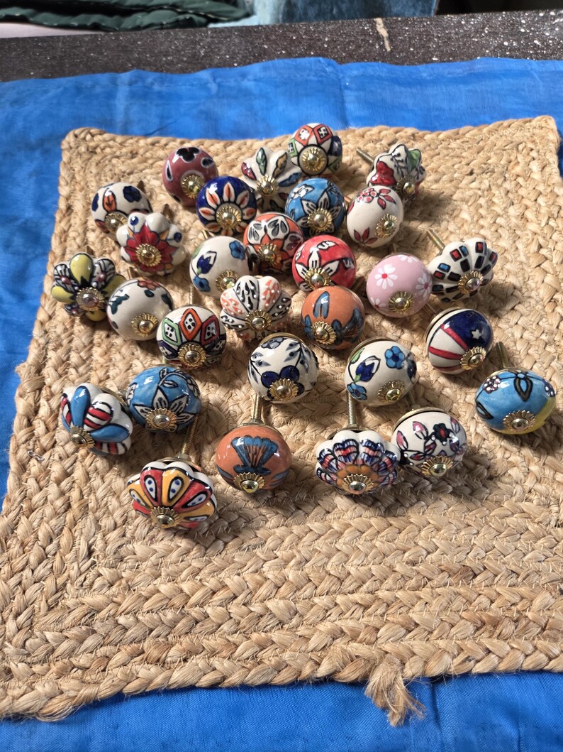 May include: A collection of twenty-four round ceramic cabinet knobs featuring various hand-painted floral and geometric patterns in colours including blue, red, orange, pink, and white, each finished with a central gold-toned metal hardware piece.