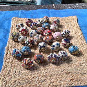 May include: A collection of round ceramic cabinet knobs featuring various colourful hand-painted floral and geometric patterns. Each knob has a central gold-toned metal hardware piece and a smooth, glossy finish with diverse designs in red, blue, yellow, and pink.