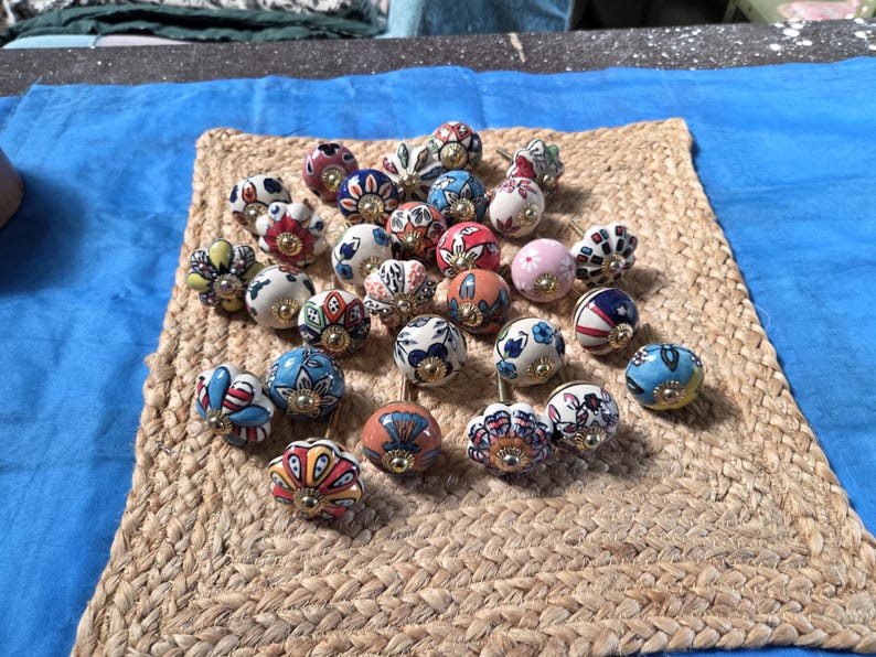 May include: A collection of round ceramic cabinet knobs featuring various colourful hand-painted floral and geometric patterns. Each knob has a central gold-toned metal hardware piece and a smooth, glossy finish with diverse designs in red, blue, yellow, and pink.