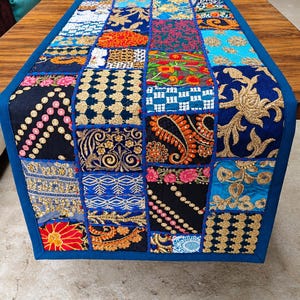 May include: A rectangular table runner featuring a patchwork design of various fabric squares. The squares display diverse patterns, including floral motifs, paisley prints, and geometric shapes in vibrant colors like blue, red, gold, and black.