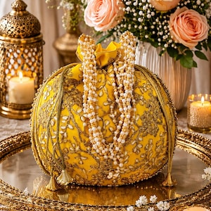 Könnte beinhalten: Ein leuchtend gelber Beutel mit Kordelzug, verziert mit aufwendigen goldenen Metallstickereien und Blumenmustern. Die Tasche ist mit langen, herabhängenden Strängen aus weißen Kunstperlen geschmückt, die als Griff oder Akzent dienen und goldene Quasten haben.