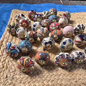 May include: A collection of round ceramic drawer knobs featuring various hand-painted floral and geometric patterns in vibrant colours including red, blue, yellow, orange, and pink, each finished with a central gold-toned metal hardware component.