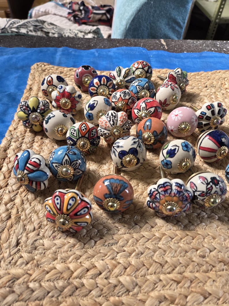 May include: A collection of round ceramic drawer knobs featuring various hand-painted floral and geometric patterns in vibrant colours including red, blue, yellow, orange, and pink, each finished with a central gold-toned metal hardware component.