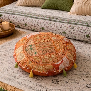 Pode incluir: Uma almofada de chão redonda com um design de patchwork laranja vibrante, bordados dourados intrincados, detalhes em fio metálico e pequenas borlas verdes presas ao perímetro. A superfície exibe vários painéis de tecido texturizado.