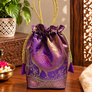 May include: A purple drawstring pouch featuring a gold peacock motif and intricate floral patterns. The bag has a gathered top, two gold twisted rope handles, and matching gold drawstring cords with purple triangular tassels at the ends.