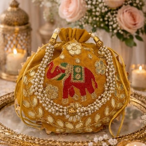 Può includere: Borsa con coulisse giallo senape con un motivo a elefante rosso ricamato. La borsa è adornata con abbellimenti dorati e un cinturino di perle. La borsa è appoggiata su uno specchio con bordo dorato, con candele e fiori sullo sfondo.