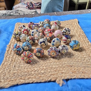May include: A collection of round ceramic cabinet knobs featuring various colourful hand-painted floral and geometric patterns. Each knob has a central gold-toned metal hardware piece and a smooth, glossy finish with diverse designs in blue, red, pink, and yellow.