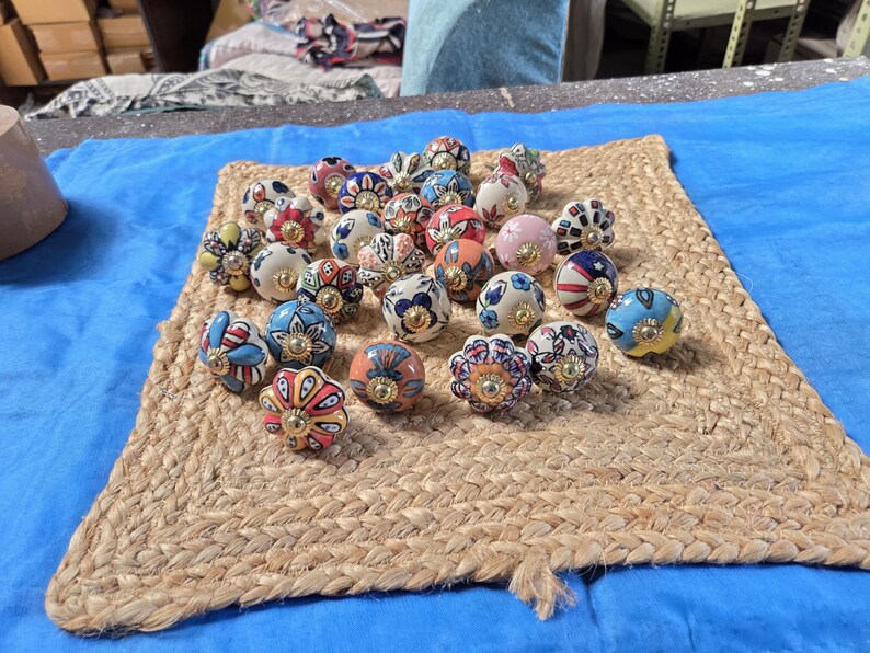 May include: A collection of round ceramic cabinet knobs featuring various colourful hand-painted floral and geometric patterns. Each knob has a central gold-toned metal hardware piece and a smooth, glossy finish with diverse designs in blue, red, pink, and yellow.
