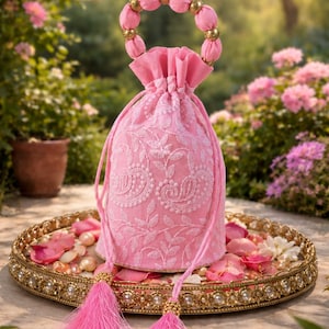 May include: A pink embroidered drawstring bag with a beaded handle and tassel accents. The bag is displayed on a gold-rimmed tray with rose petals and pearls. The bag's design features floral embroidery.