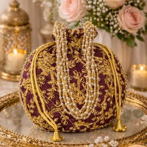 May include: A deep purple drawstring pouch featuring intricate gold thread embroidery and a decorative handle made of multiple strands of small white beads. The bag has a rounded shape with gold cord drawstrings that end in small metallic tassels.