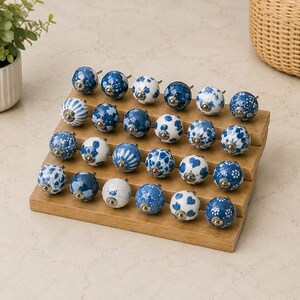 Pu&ograve; includere: Una collezione di ventiquattro pomelli rotondi in ceramica per mobili disposti su un espositore in legno a gradini. Ogni pomello presenta un motivo unico in blu e bianco, inclusi disegni floreali, a righe e a pois, con una base in metallo argentato.