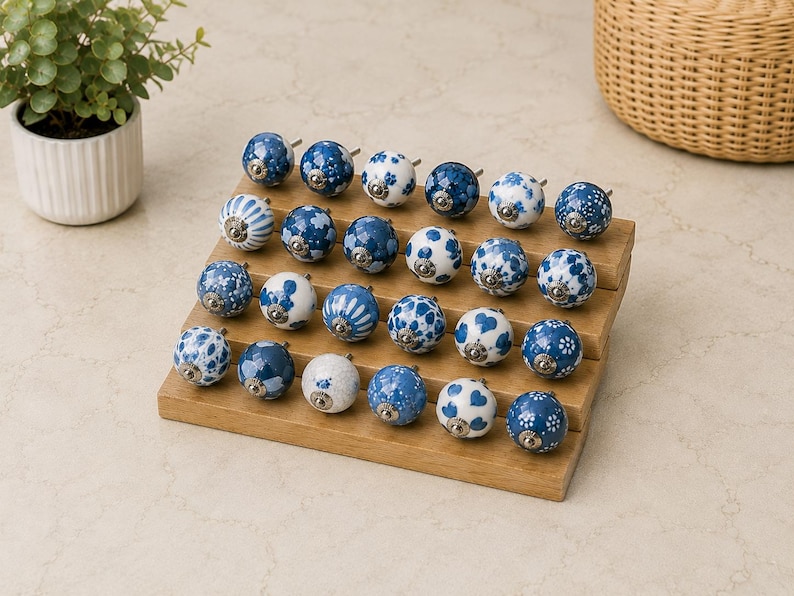 Pu&ograve; includere: Una collezione di ventiquattro pomelli rotondi in ceramica per mobili disposti su un espositore in legno a gradini. Ogni pomello presenta un motivo unico in blu e bianco, inclusi disegni floreali, a righe e a pois, con una base in metallo argentato.