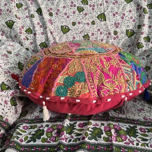 May include: A round, multi-colored patchwork floor cushion featuring intricate embroidery, sequins, and beadwork in shades of pink, orange, green, and blue. The cushion has a solid dark pink side panel with small white tassels attached along the edge.