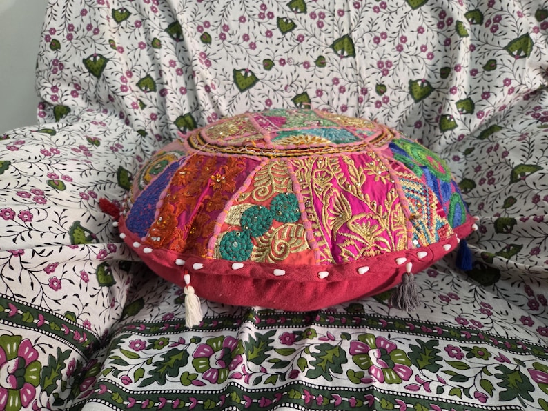 May include: A round, multi-colored patchwork floor cushion featuring intricate embroidery, sequins, and beadwork in shades of pink, orange, green, and blue. The cushion has a solid dark pink side panel with small white tassels attached along the edge.