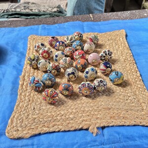 May include: A collection of round ceramic cabinet knobs featuring various colourful hand-painted floral and geometric patterns. Each knob is finished with a central gold-toned metal hardware piece, showcasing a diverse array of vibrant colours.