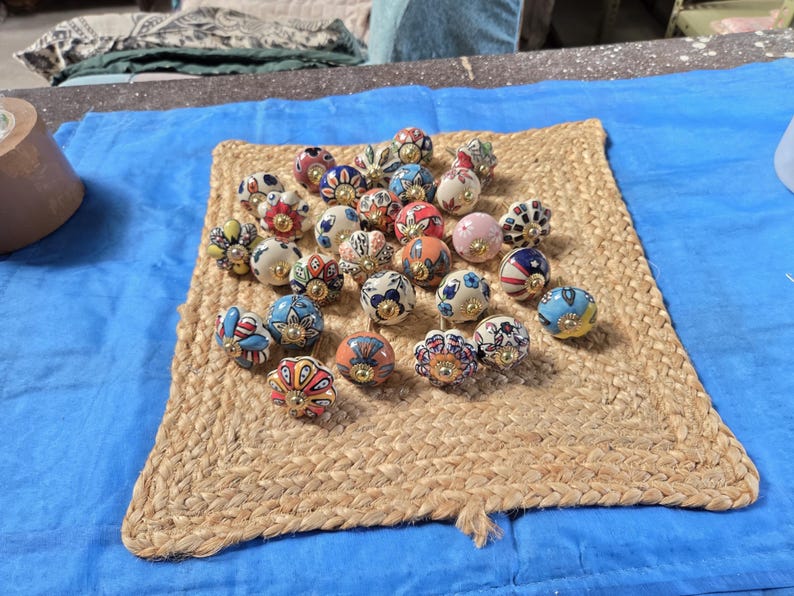 May include: A collection of round ceramic cabinet knobs featuring various colourful hand-painted floral and geometric patterns. Each knob is finished with a central gold-toned metal hardware piece, showcasing a diverse array of vibrant colours.