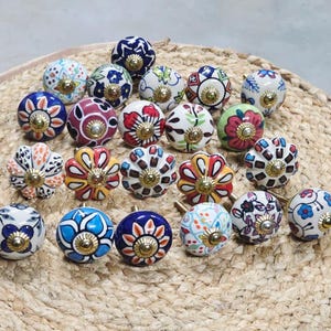 Puede incluir: Una colección de pomos decorativos de cerámica con diseños florales y geométricos. Los pomos son redondos, con una variedad de colores que incluyen azul, rojo, amarillo y blanco. Cada pomo tiene una base de metal dorado y está dispuesto sobre una estera tejida.